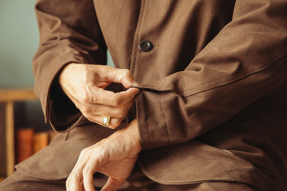 Image of model wearing Conkers Bruton Blazer in dark brown with hands messing with the sleeve buttons on jacket.