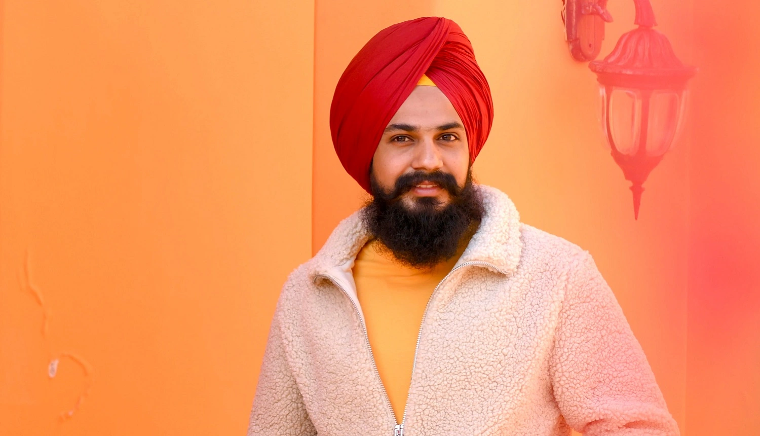 Em artigo sobre falhas na barba, imagem mostra um indiano sikh, com barba escura bastante longa, casaco branco, camisa amarela e turbante vermelho, em frente a parede laranja.