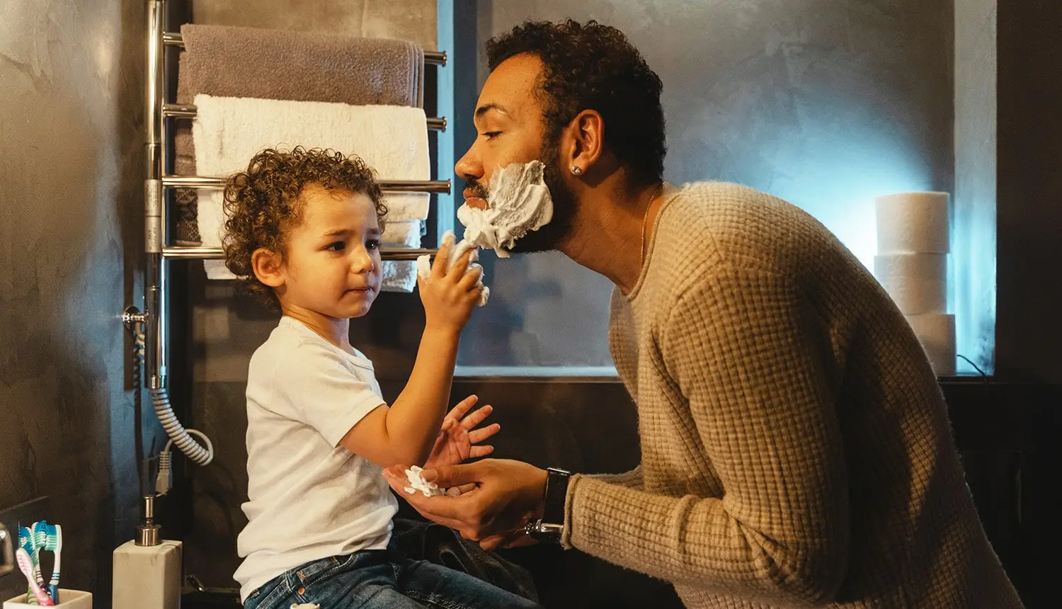 Em banheiro, pai de pele parda, barba, cabelos cacheados curtos e blusa bege, barbado, faz a barba com seu filho criança, de camiseta branca e calça jeans. O filho passa espuma de barbear no rosto do pai.
