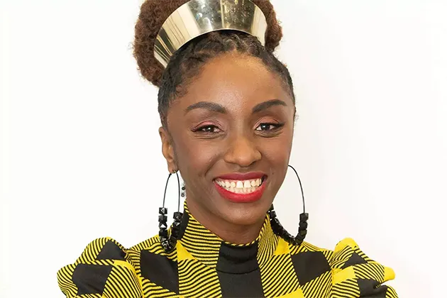 Foto de Nina Silva, que usa uma tiara e cabelo em trança, grandes brincos e blusa quadriculada xadrez em amarelo e preto. Ela sorri para a câmera.