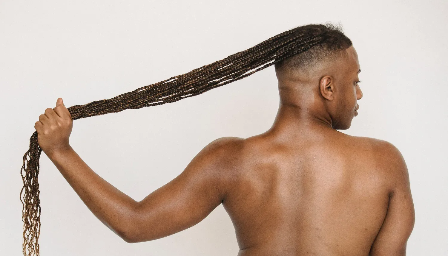 Foto de homem negro de costas, com cabelo raspado nas laterais e parte posterior da cabeça, sem camisa e longas tranças afro. As tranças partem do alto da cabeça e ele segura a parte inferior delas com a mão esquerda. A foto ilustra a força do cabelo, com o uso do Capalgin em shampoos antiqueda.