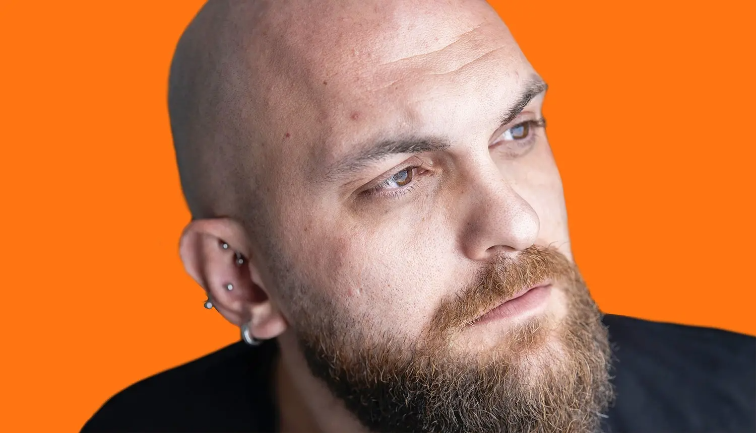 Em fundo laranja, foto de homem olhando para a esquerda, piercings na orelha e careca. Ele tem a barba longa, entre ruiva e castanho-clara, e olhos castanhos. Usa camiseta cinza e escura.