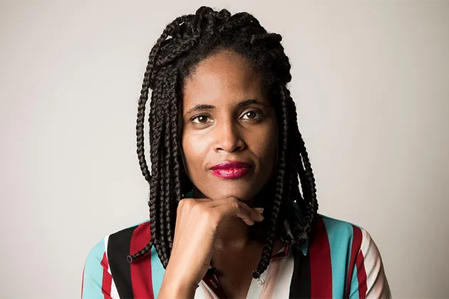 Foto de Djamila Ribeiro, que é mulher negra, usando tranças em cabelo comprido, batom rosa e camisa listrada preta, branca, azul e vermelha.