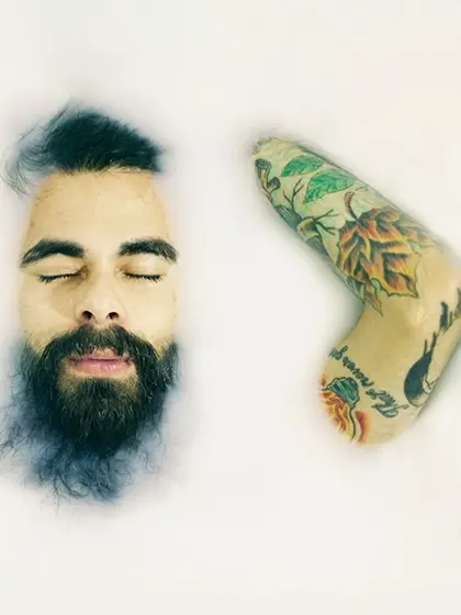 Em artigo sobre autocuidado para portadores de HIV e aids, foto mostra homem branco com barba mergulhado em líquido branco espesso. Vê-se seu rosto e o braço esquerdo, que tem tatuagens.