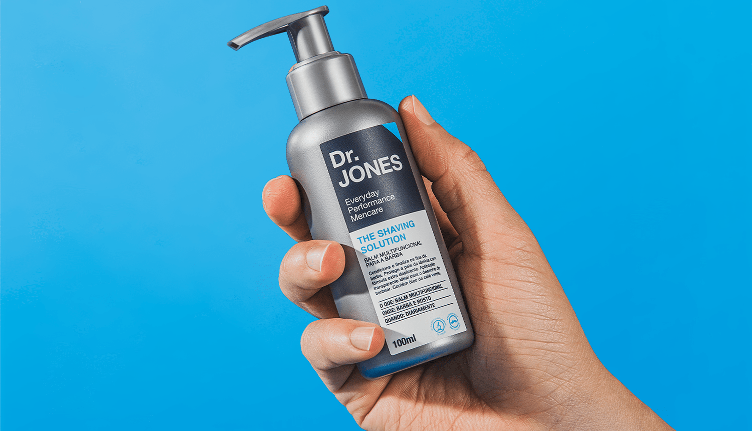 Em fundo azul, foto mostra mão segurando embalagem do balm para barbear THE SHAVING SOLUTION, da Dr. JONES, que contém ativo extraído da raiz de gengibre.