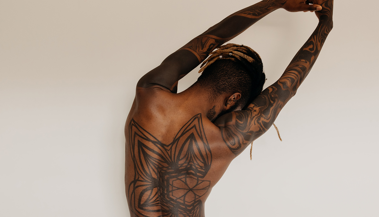 Homem negro, cabelo raspado na lateral, com tranças descoloridas. Ele está de costas, que são tatuadas, alongando-as, em frente a um fundo de cor clara.