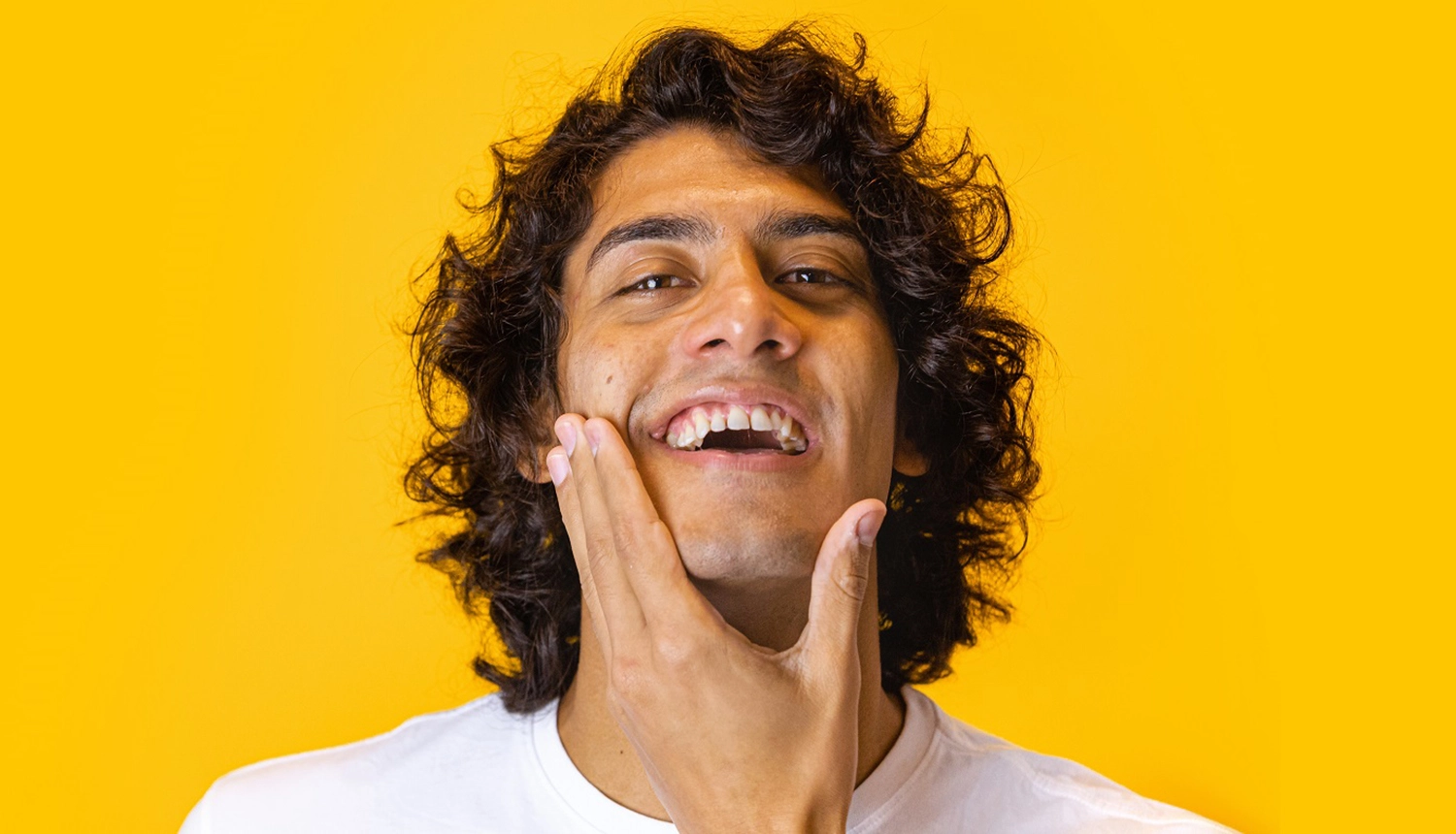 Em artigo sobre skincare (cuidados com a pele masculina), imagem em fundo amarelo traz homem de pele parda, olhos castanhos e cabelo cacheado médio e castanho sorrindo, com as mãos no queixo.
