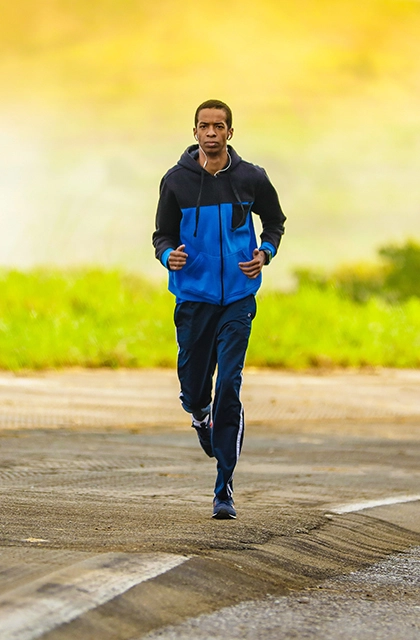 Em artigo sobre autocuidado para portadores de HIV e aids, foto mostra homem negro correndo sobre uma pista e vestindo agasalho azul e calça azul. Ao fundo, há paisagem com mata.