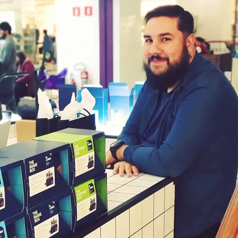 Usando uma camisa social azul, Rafael Rosa, que tem pele clara, barba grande e cabelos curtos castanhos, aparece sentado em um estande da Dr. JONES, ao lado de kits da marca.