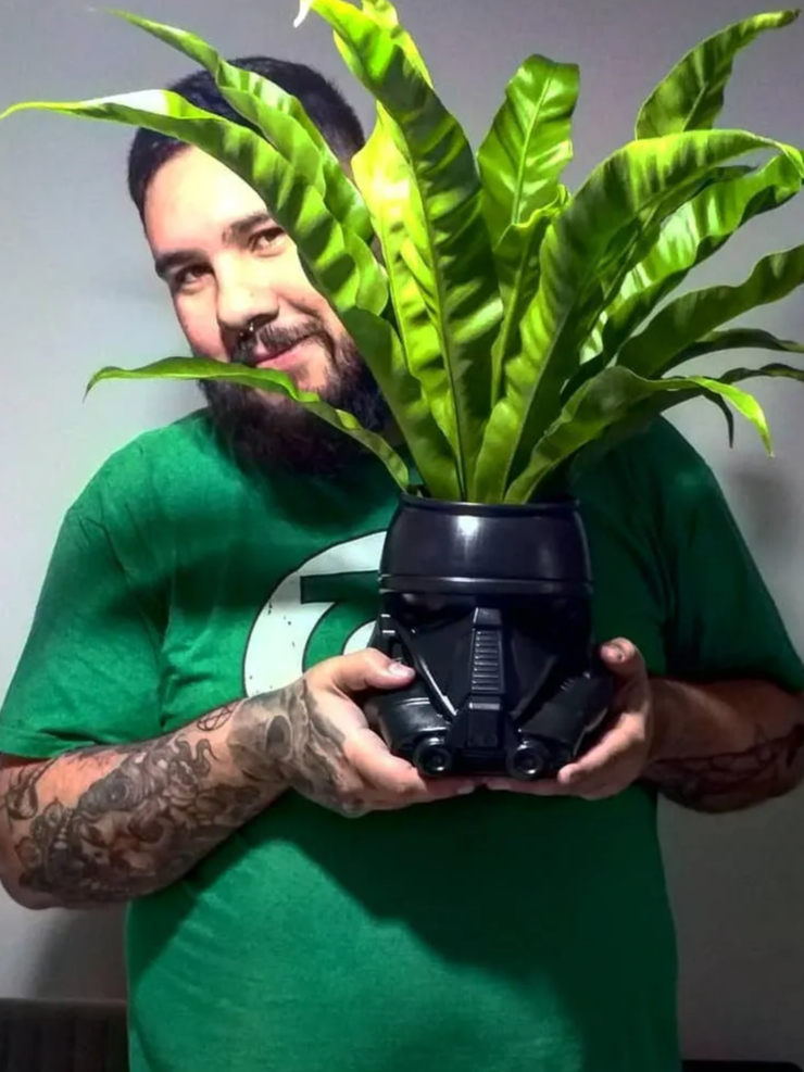 Foto de Rafael Rosa, da Dr. JONES, segurando um vaso com planta. O vaso tem o design de um soldado de Star Wars, preto, e Rafael usa uma camiseta com o logo do herói Lanterna Verde.