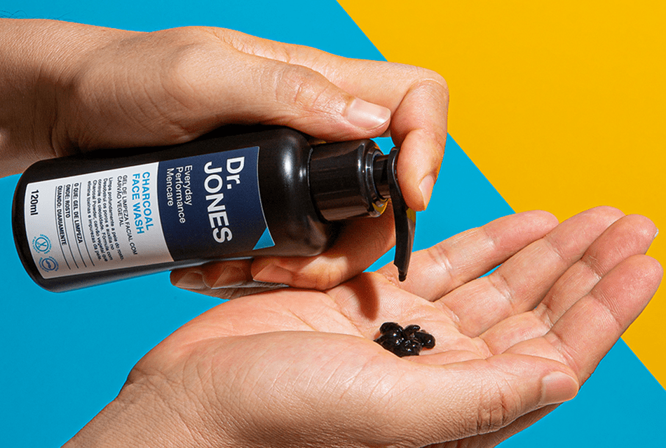 Foto do produto CHARCOAL FACE WASH, da Dr. JONES. Uma pessoa pressiona com a mão direita o produto, que derrama em pequena quantidade na outra mão aberta, em fundo bicolor azul e amarelo.