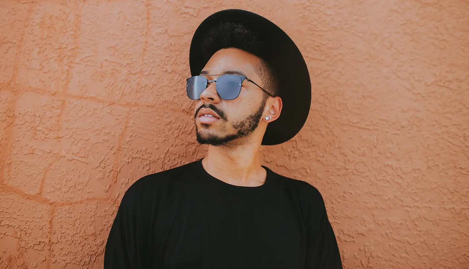 Foto de homem pardo, com barba aparada, cabelos e barba escuros, usando chapéu e camiseta preta em fundo laranja. A barba é fina nas laterais, com bigode e cavanhaque.