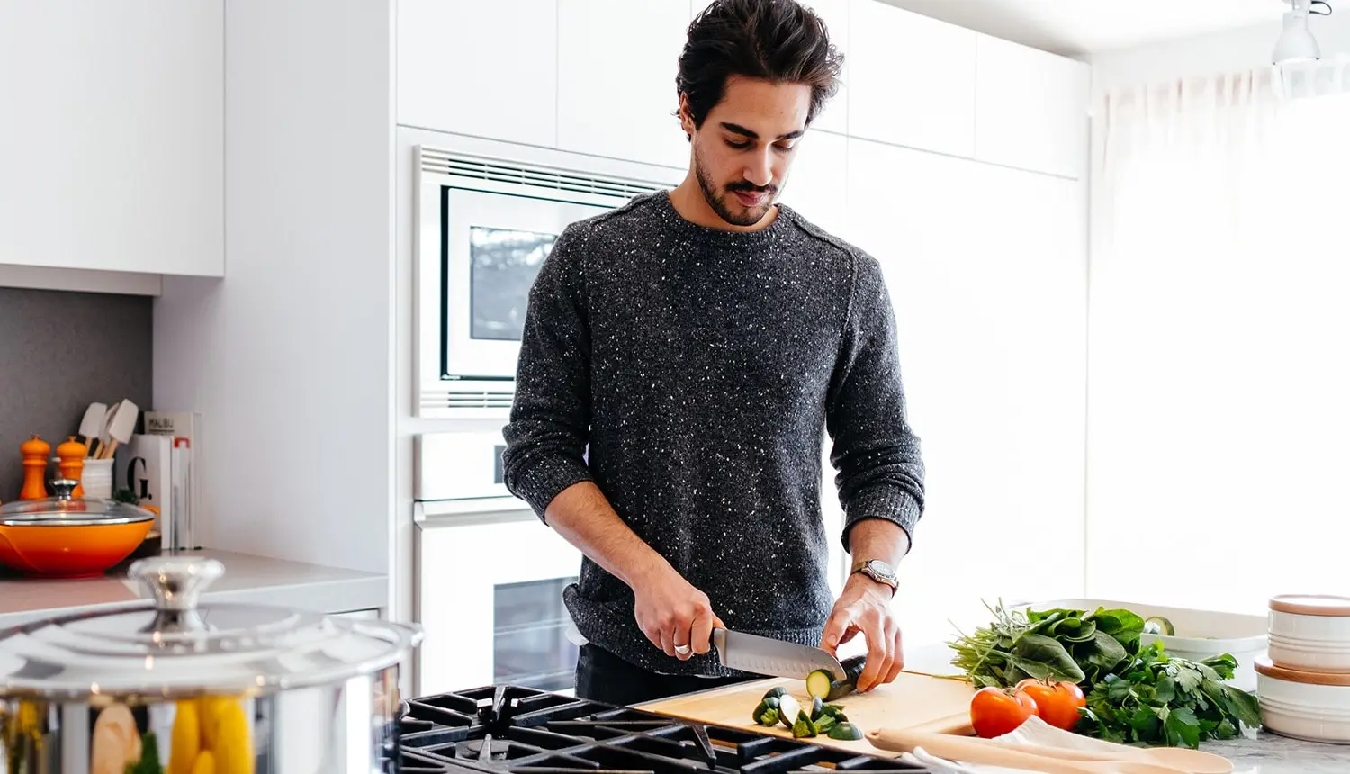 Homem de pele clara, cabelos curtos e densos, castanhos, bigode e cavanhaque e blusa cinza prepara alimentos vegetais em cozinha: folhas escuras, tomates e pepinos, que ele corta com a faca. À esquerda, encontram-se armário e panela.