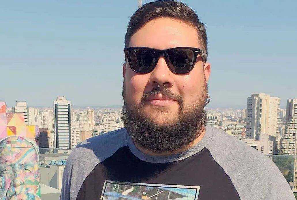 Foto de Rafae Rosa, da Dr. JONES, usando óculos escuros e camiseta preta de mangas cinza em espaço aberto de prédio em São Paulo, com a imagem da cidade ao fundo.