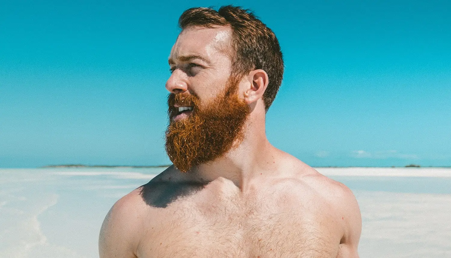 Em frente a praia deserta com muito sol, homem sem camisa e de pele clara, ruivo, olha para a esquerda. Ele tem a barba comprida, em alusão aos cuidados necessários para o verão.