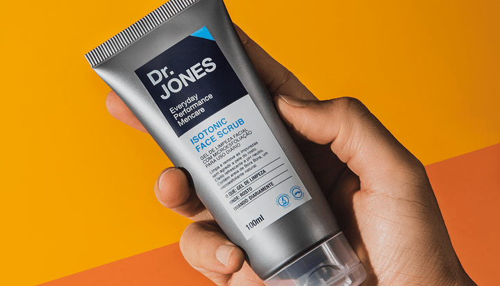 Em fundo laranja e marrom-claro, foto mostra uma mão de pele clara segurando a embalagem do esfoliante Isotonic Face Scrub, da Dr. JONES.