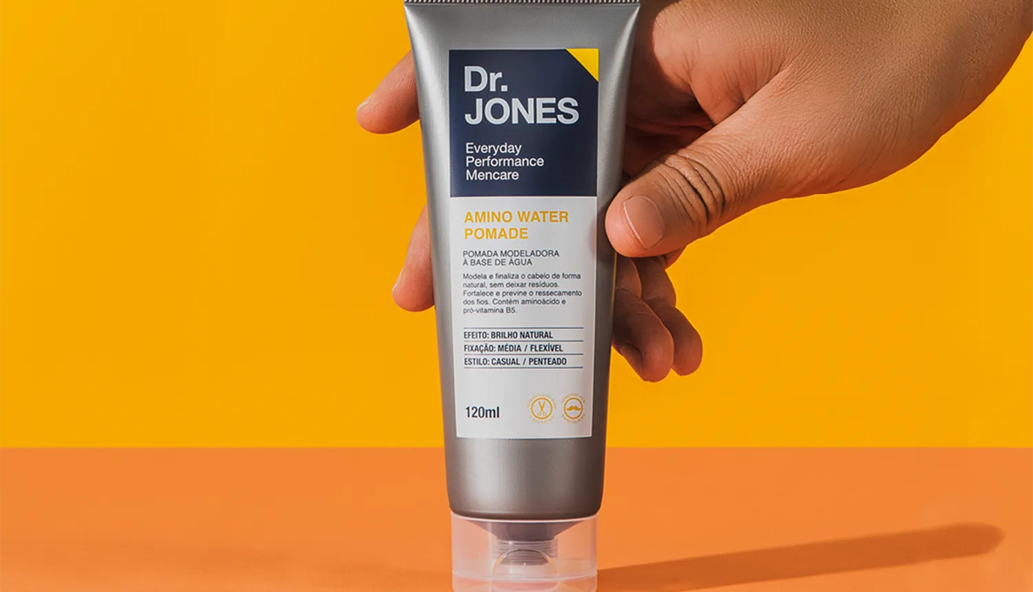 Em fundo amarelo com mesa laranja, foto da Amino Water Pomade, pomada de cabelo da Dr. JONES, para cabelos masculinos, que utiliza Carbocisteína na fórmula. A embalagem cinza é segurada por uma mão.