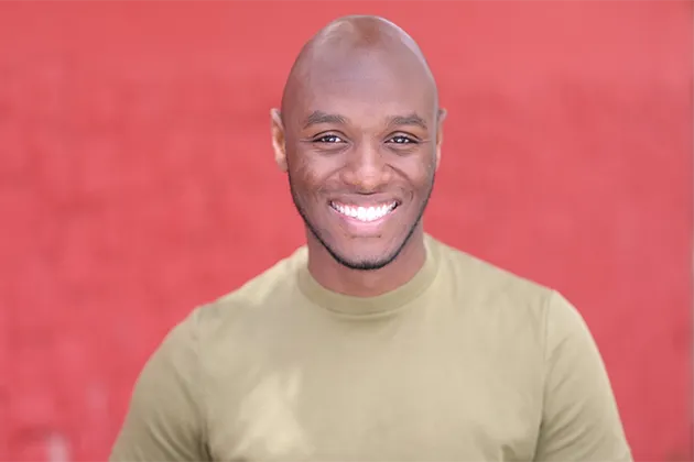 Foto de AD Junior, que é um homem negro, sem cabelos, sorrindo para a câmera, camisa bege-esverdeada e barba, sem bigode ou cavanhaque.