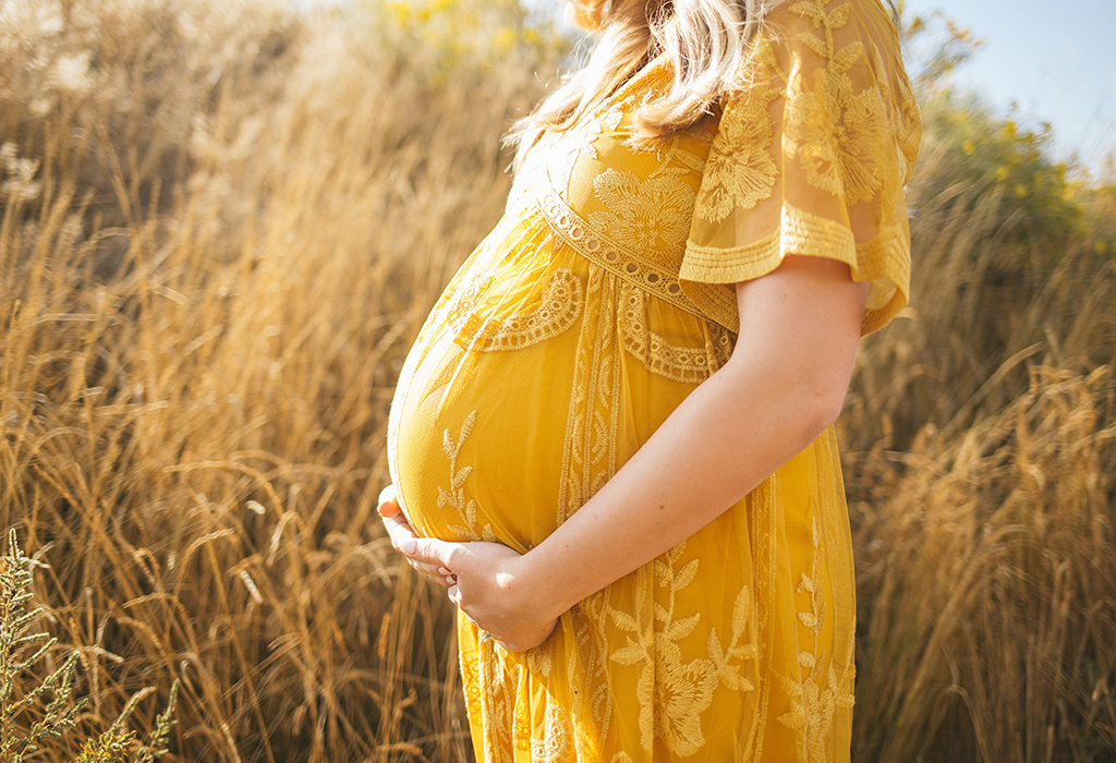 Eine schwangere Frau im gelben Kleid steht vor einem Getreidefeld und umfasst ihren Bauch.