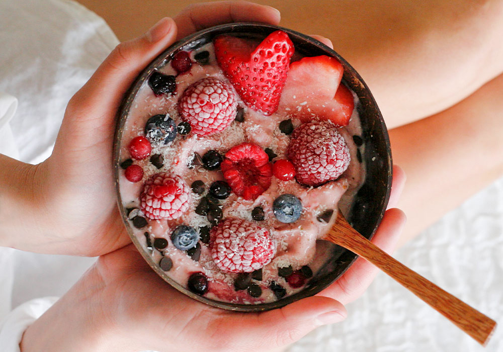 2 Frauenhände halten eine Smoothiebowl, dekoriert mit Beeren und Kakaonibs