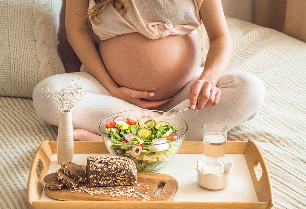 Eine schwangere Frau sitzt im Schneidersitz auf einem Bett und legt eine Hand auf ihren nackten Bauch, mit der anderen Hand hält sie eine Gabel über die vor ihr stehende Schüssel mit Salat.