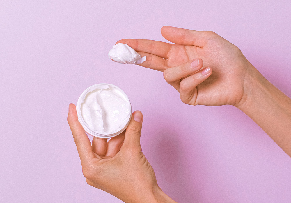 Neben einer geöffneten Dose mit Creme ist eine Hand mit Creme auf den Fingern.