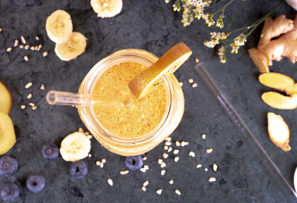 Ein Glas mit gelbem Smoothie steht auf einer grauen Fläche, daneben liegen Ingwer und Gewürze.