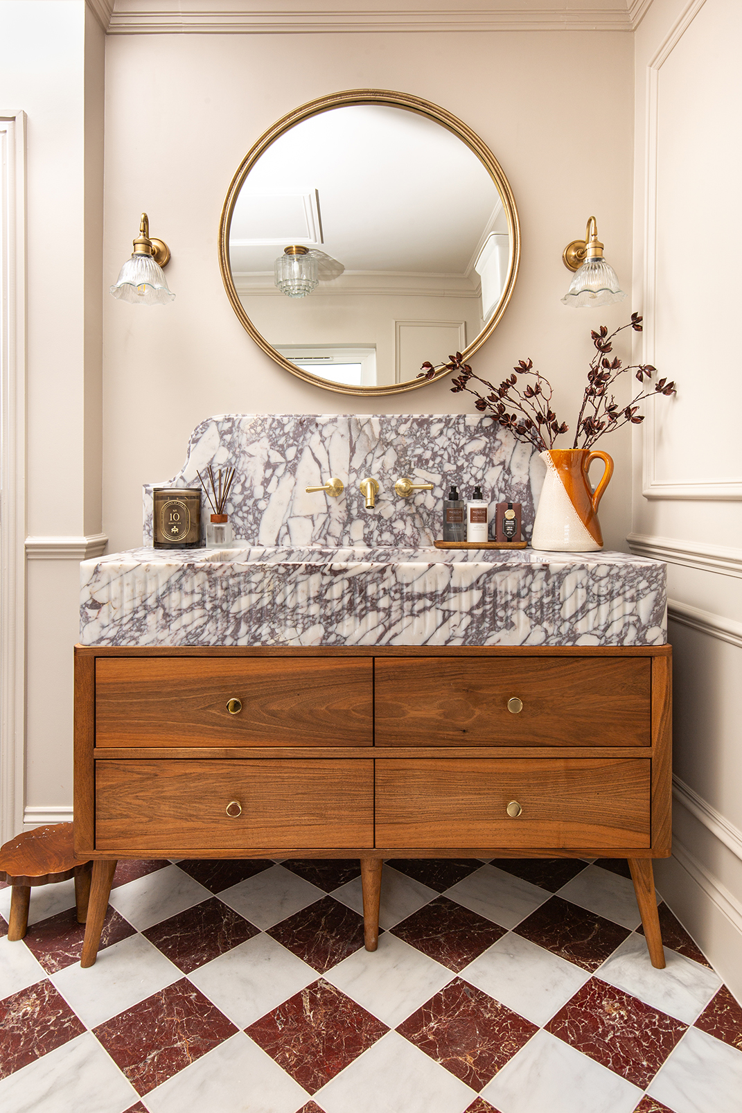 Mid-century bathroom with a marble sink, circular mirror, and wooden vanity with chequered Riviera Burgundy & White. Warm lighting from brass sconces creates a cozy, elegant ambiance.