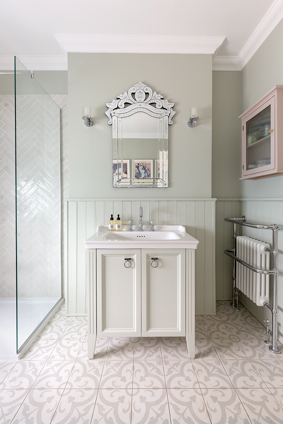 Elegant bathroom with a vintage vibe, featuring a decorative mirror above a white vanity, patterned Clarence Mews tiles, glass shower, and light green walls.
