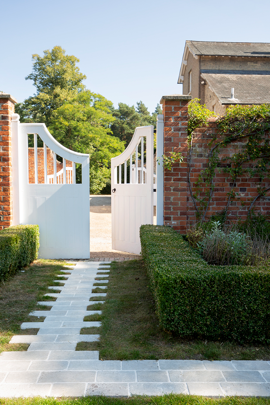 A white garden gate is open on a sunny day, revealing a brick Clermont Gris Aged tumbled cobble path flanked by manicured hedges. The scene feels inviting and peaceful.