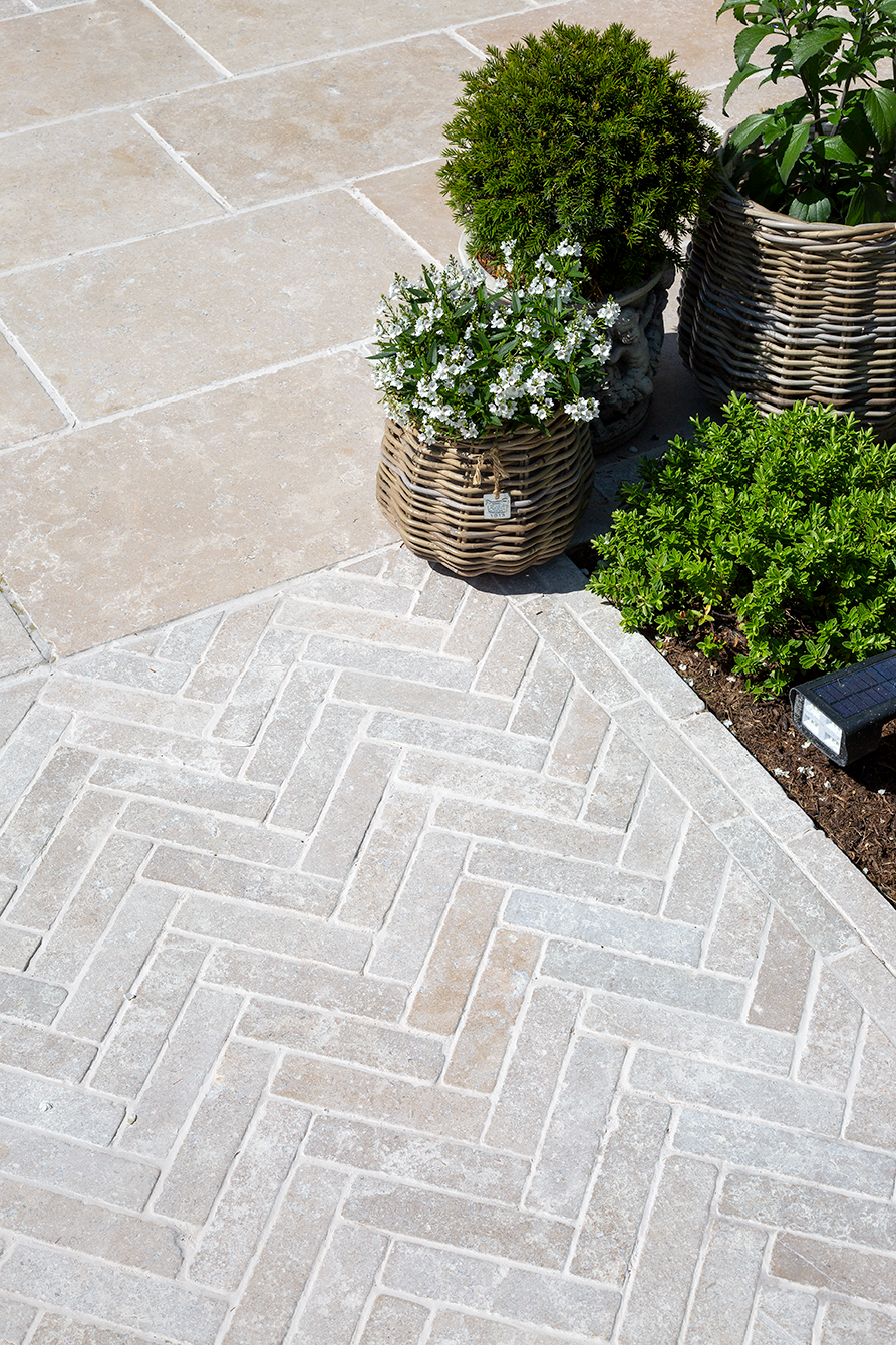 A Dijon TUmbled Limestone patio with light beige tiles; a herringbone pattern contrasts with larger tiles. Nearby are wicker baskets with lush green plants and white flowers.