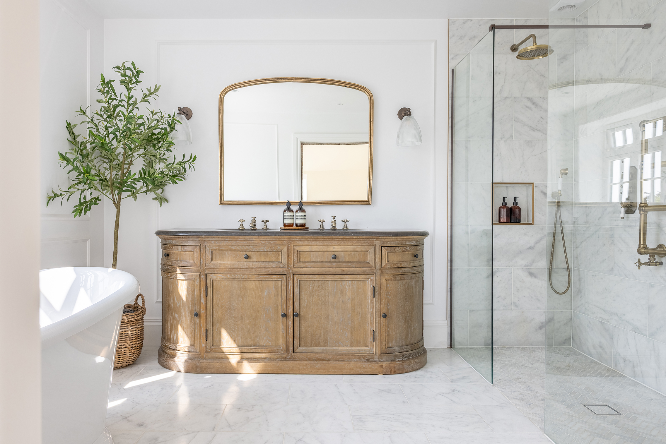 Elegant bathroom with Carrara Bianco marble tiles with a rustic wooden vanity, large mirror, marble shower, and bright window. A potted plant and basket add natural elements. Serene ambiance.
