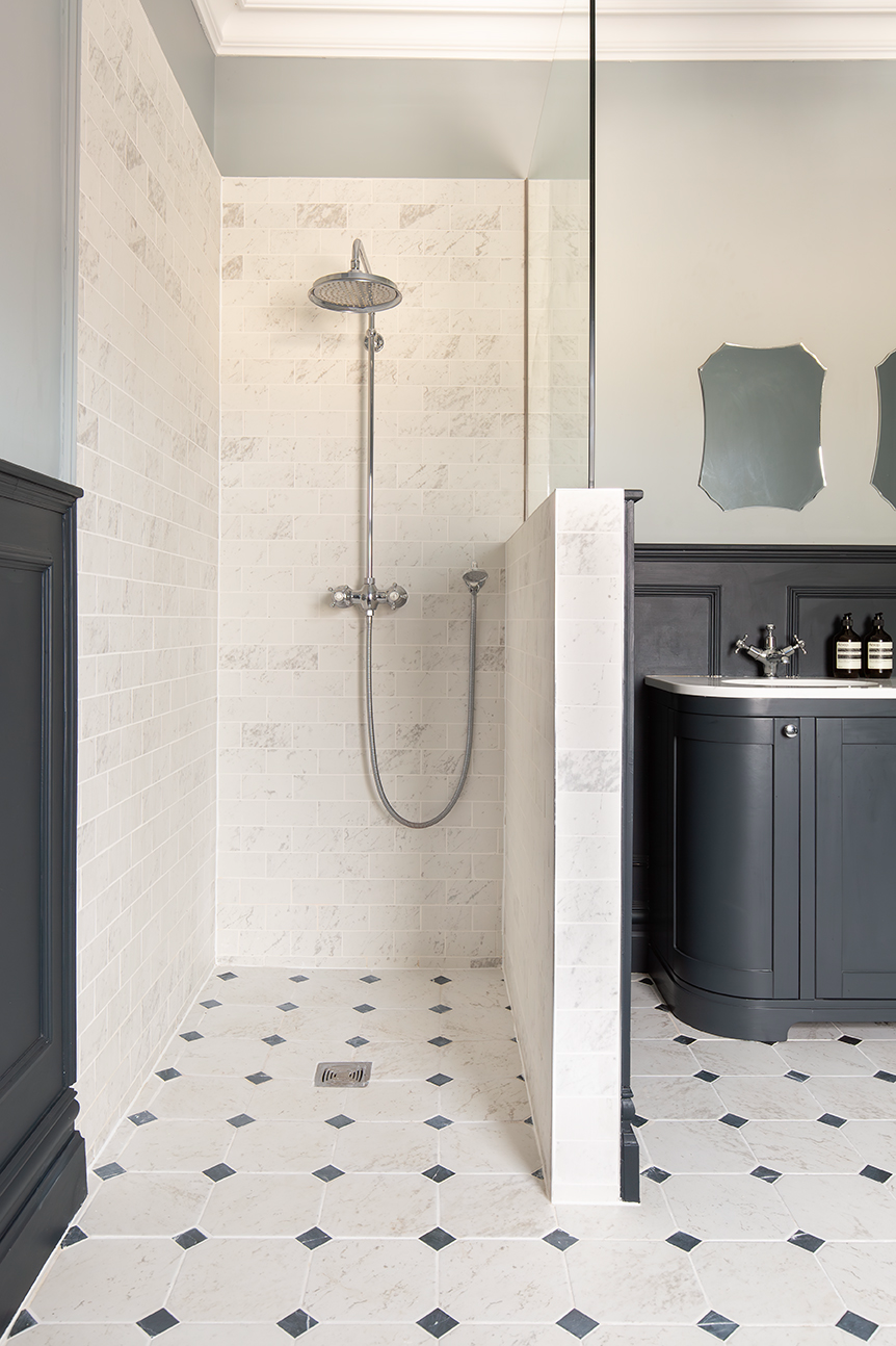 A bright bathroom with a shower featuring white marble metro tiles. The floor is tiled with limestone tiles and a dark grey vanity unit.