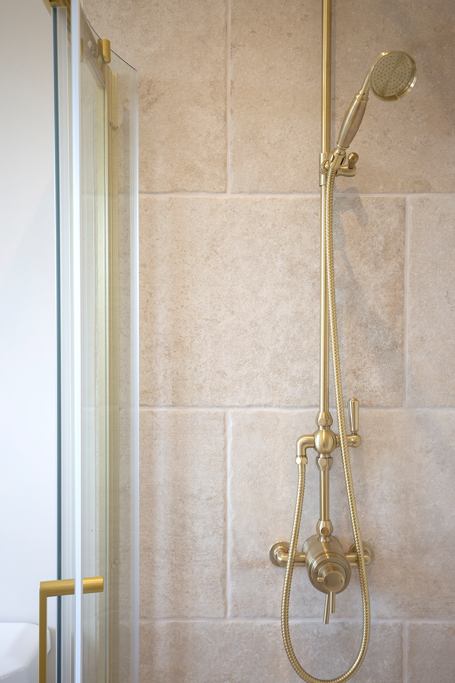 Elegant bathroom with gold shower fixtures against Chastleton Ivory stone effect tiles, next to a glass shower door. The design exudes luxury and sophistication.