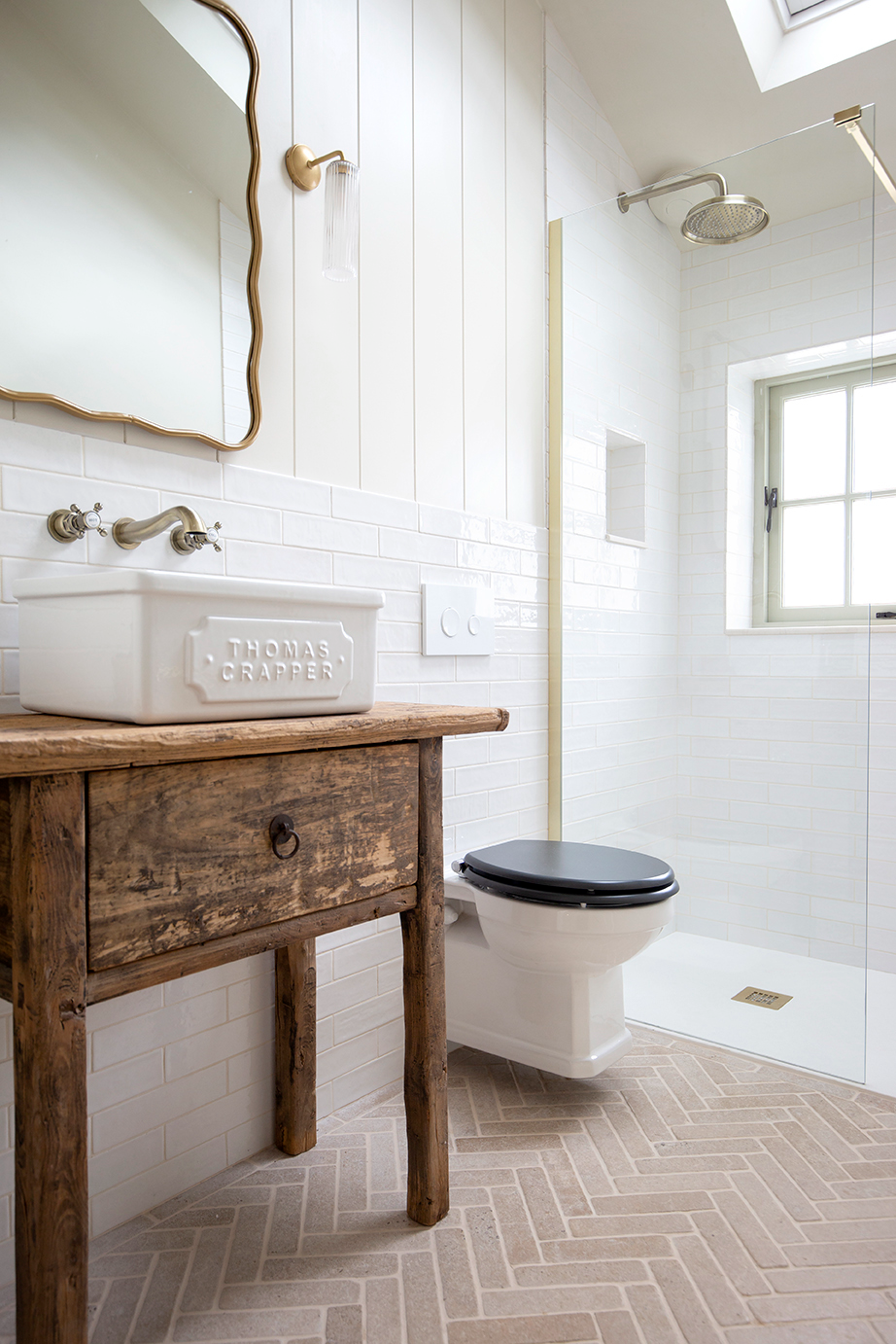 Rustic bathroom with wooden vanity, white sink, and brass fixtures. White subway tile walls, glass shower, and Tuscany Tumbled herringbone tile floor add elegance.