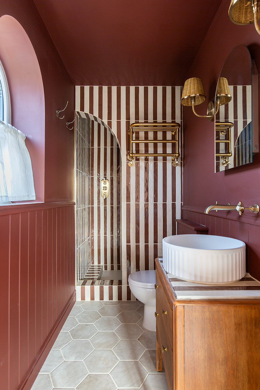 Narrow bathroom with rich maroon walls, hexagonal floor tiles, striped Luella shower tiles, round vessel sink, gold fixtures, and arched window. Elegant and warm.