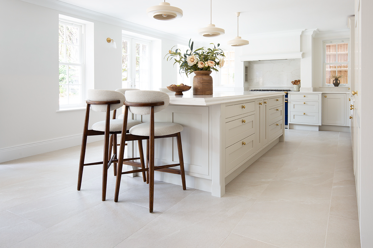 A light kitchen with white cabinets and a large island. Featuring dark wooden bar stools and a lightly coloured tiled floor.