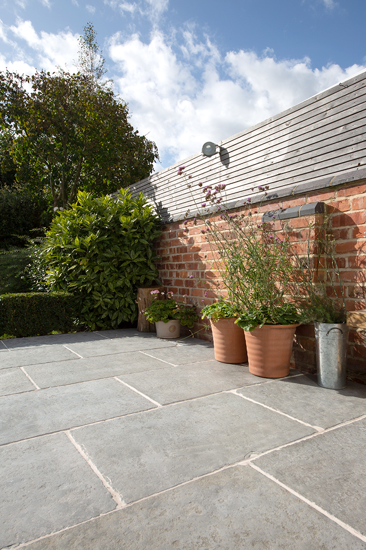 Sunny Worn Grey Antiqued patio with gray stone tiles, bordered by a red brick wall. Pots of greenery and foliage add a touch of nature. Blue sky and clouds overhead.