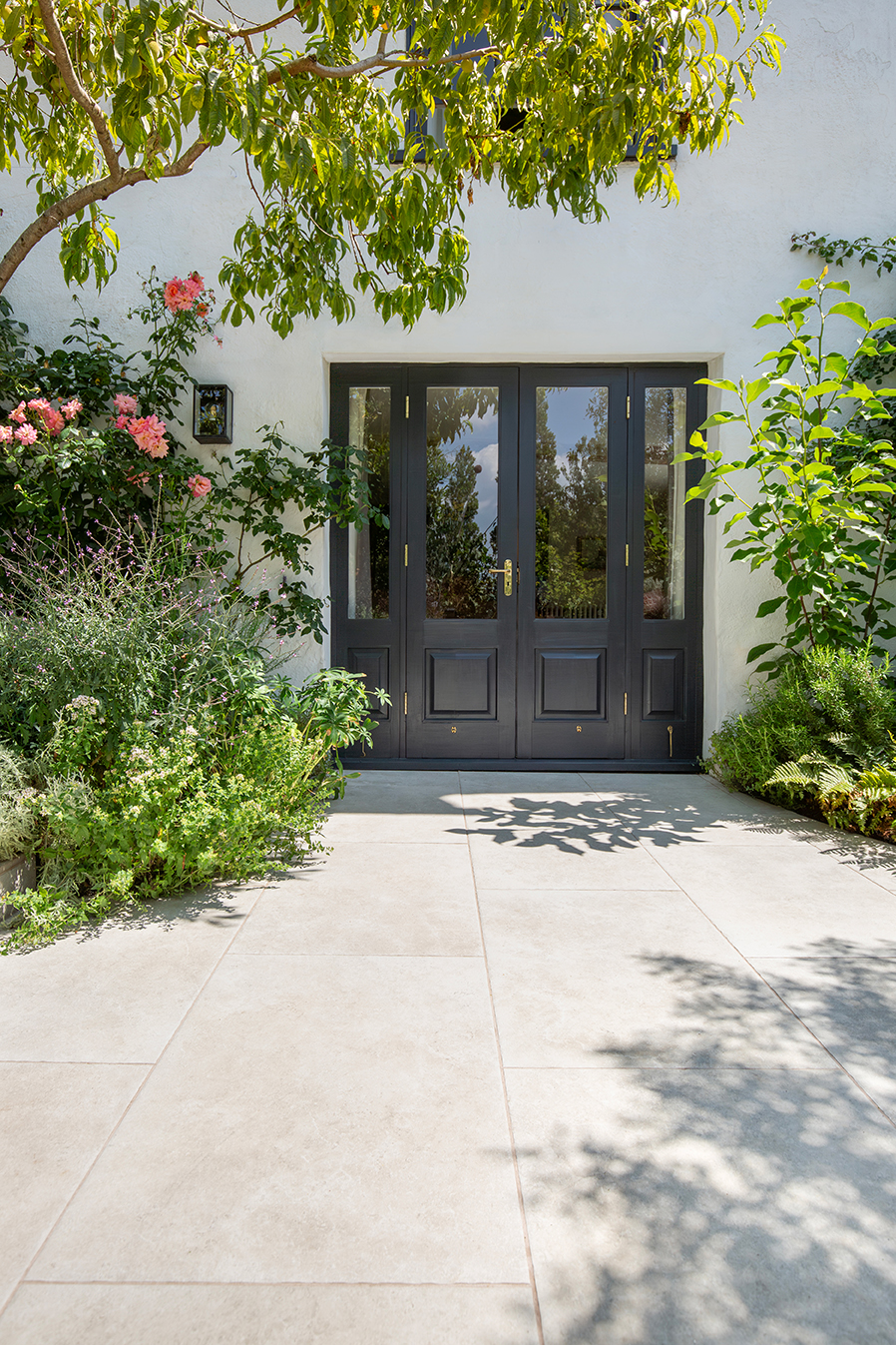 A stylish black double door with glass panels, framed by lush greenery and vibrant flowers, opens onto a sunlit Hambleton Ivory patio .