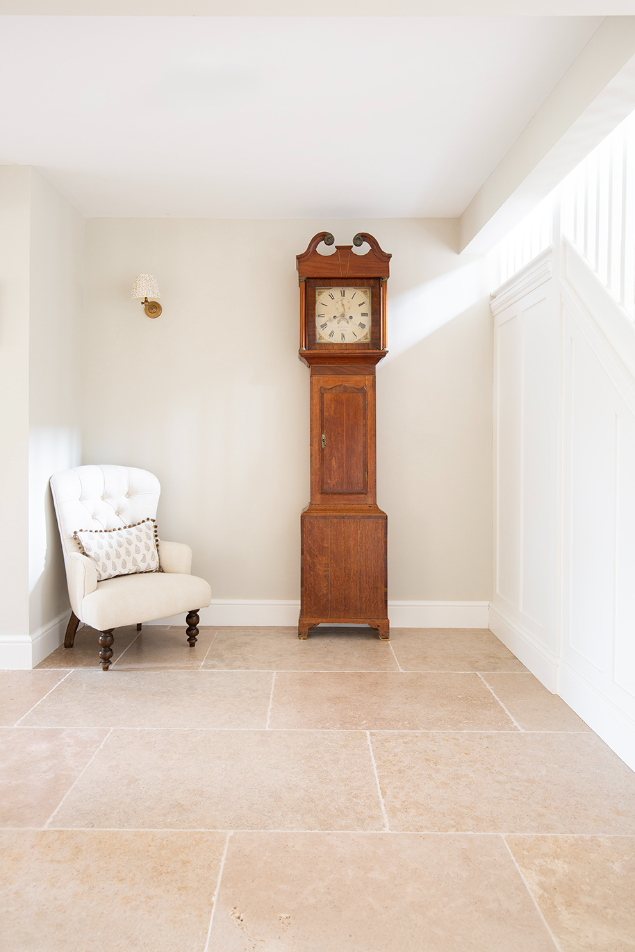 A bright, neutral hallway featuring beige limestone tiles, a white small armchair and a large wooden grandfather clock.