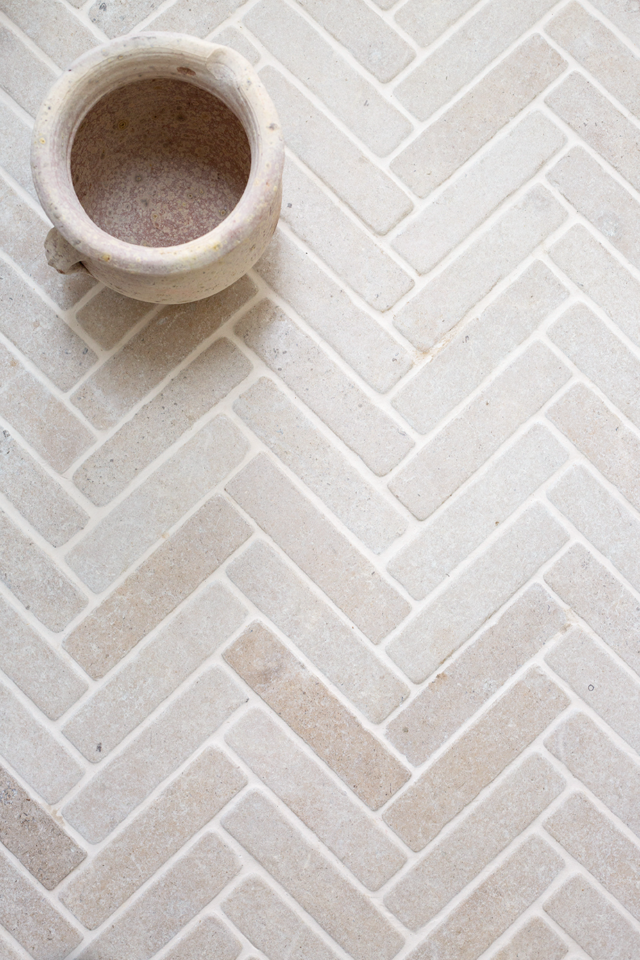 A beige ceramic pot sits on a Tuscany Tumbled herringbone-patterned tile floor. The neutral tones evoke a calm and minimalist aesthetic.
