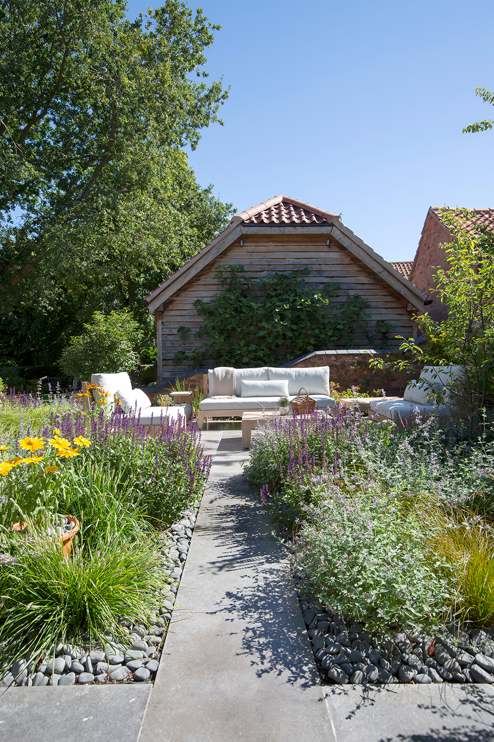 A serene garden with a paved Farrow Grey Tumbled Limestone path leading to a cozy seating area with white cushions. Vibrant flowers and greenery surround the space, under a clear blue sky.