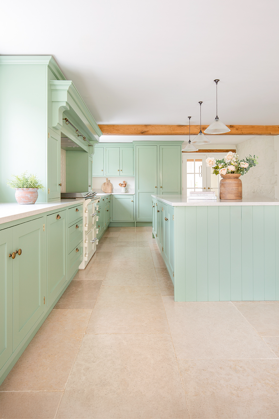 A large French inspired green kitchen with a beige limestone tile floor and large island with pendant lights hanging from the ceiling.