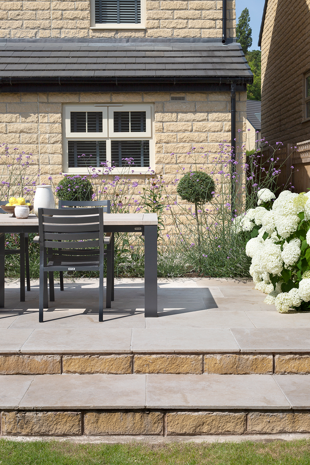 Tuscant Tumbled Limestone patio with a modern dining set on stepped tiles, bordered by lush white flowers and lavender. A beige brick house with window shutters is in the background.