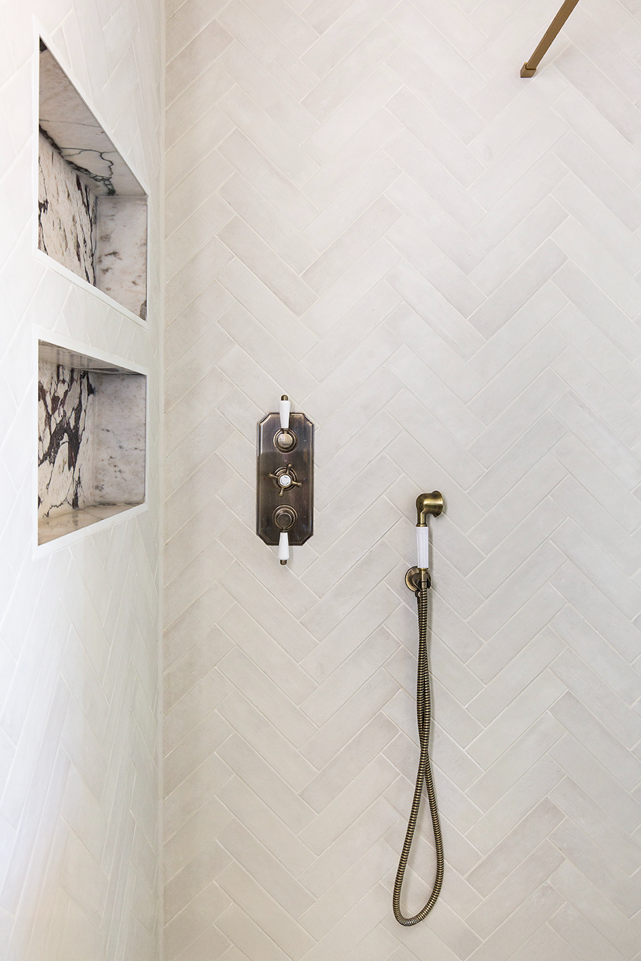 Elegant shower with diagonal white Pottery Chalk metro herringbone tiles, bronze fixtures, and a handheld showerhead. Two marble-lined niches add luxury and style.