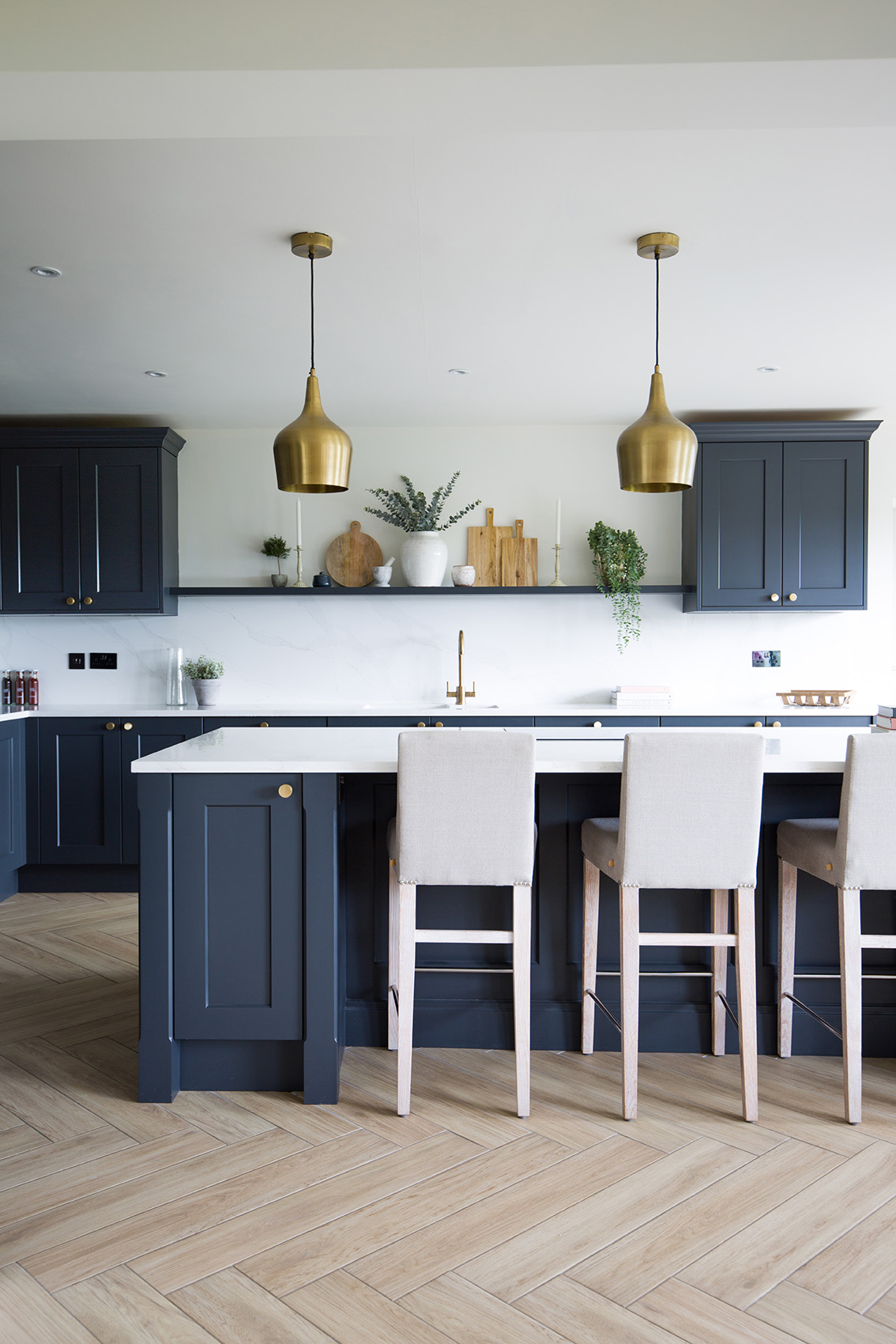 Falmouth Sandy Oak wood effect planks in a herringbone floor pattern with navy blue units in a kitchen with shelf and gold ceiling lights
