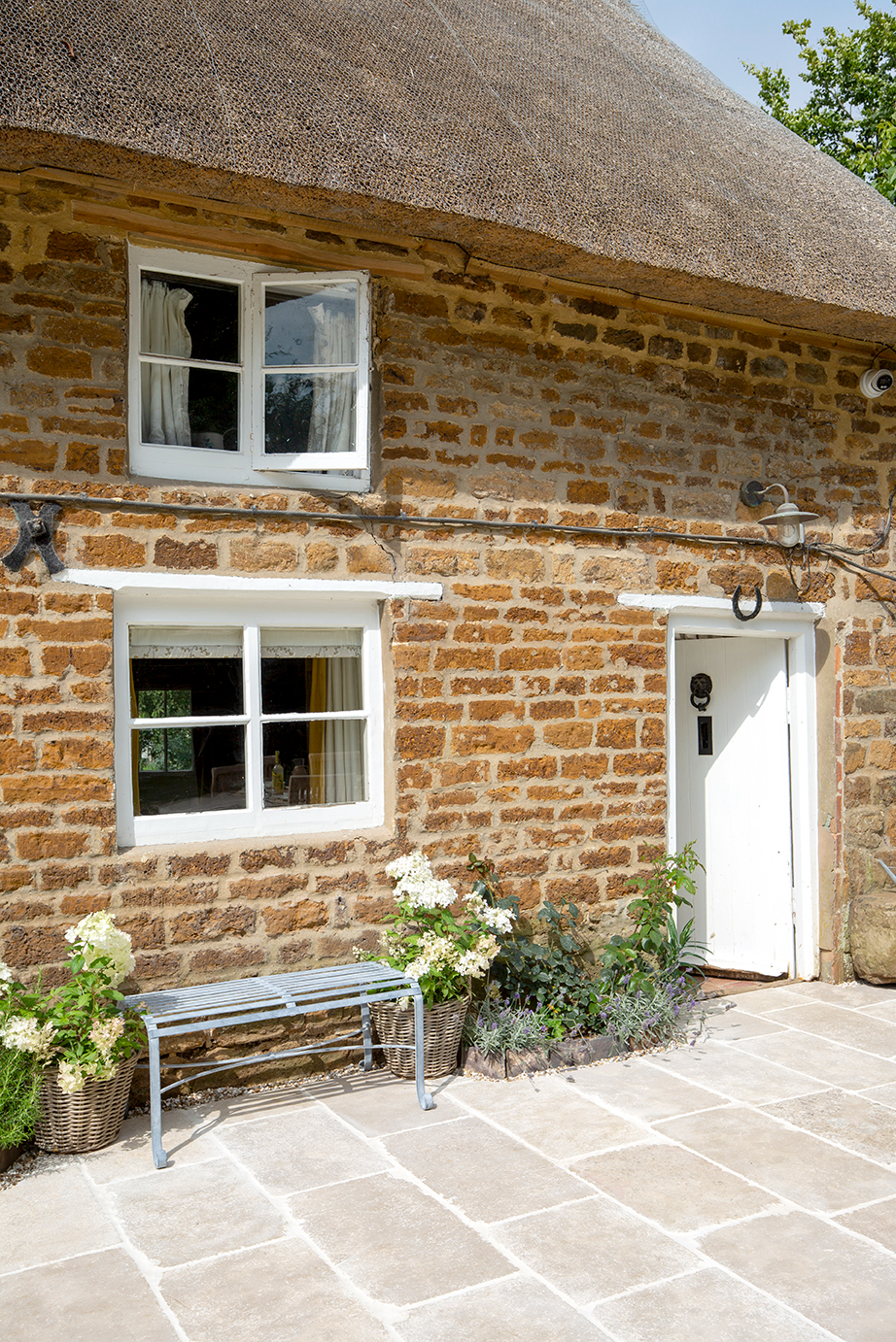 Charming stone cottage with white-framed windows, a rustic thatched roof, and a wooden door. A bench and potted plants adorn the sunlit SOrrento Aged Tumbled patio.