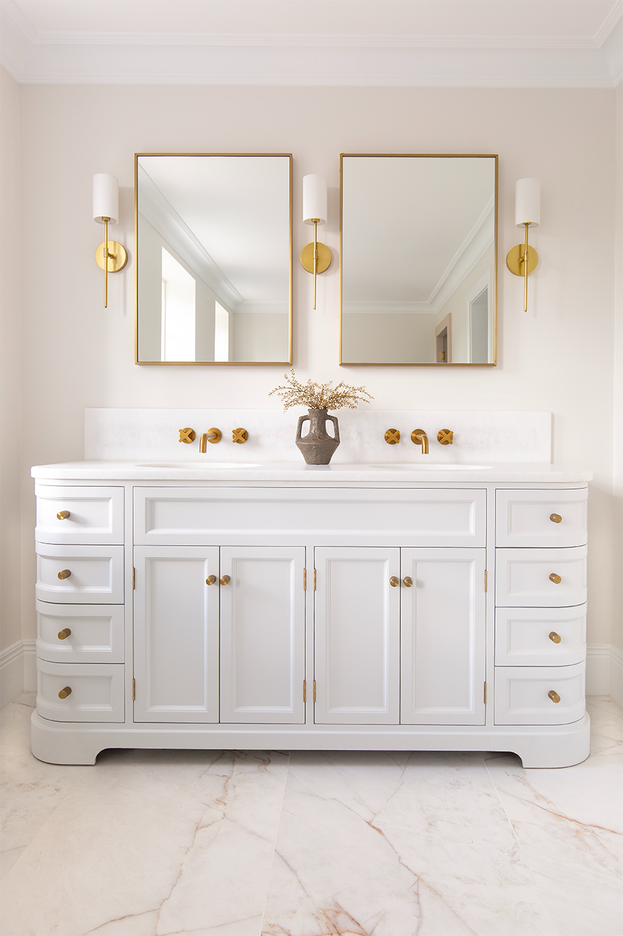 A traditional white cabinet with two mirrors and gold wall lights. Featuring white toned marble floor with pink tones.