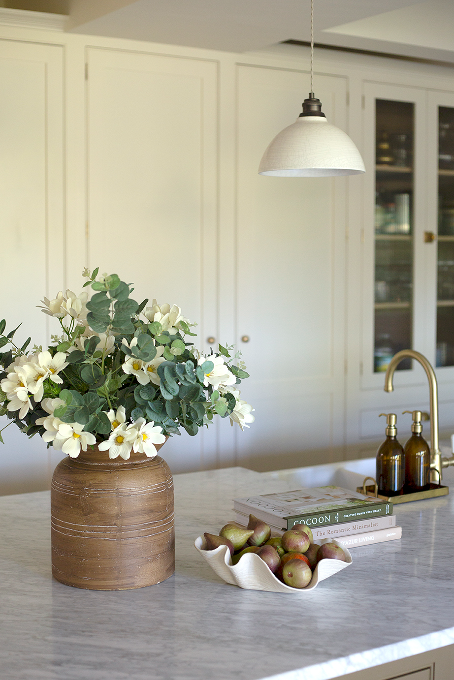 A kitchen countertop with a vase of white flowers, a bowl of pears, stacked books, a gold faucet, and a pendant light overhead.