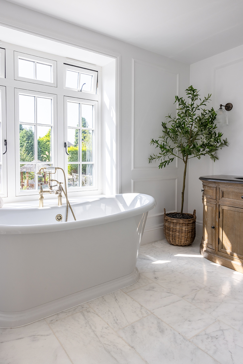Bright bathroom with Carrara Bianco Marbel with a white freestanding tub, brass faucet, large window, and a potted plant. Wooden cabinet adds a rustic touch. Minimalist and serene.